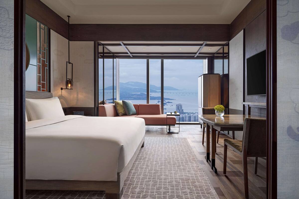 Una habitación de hotel moderna con una cama grande, un sofá, una mesa de comedor y un televisor. La habitación tiene ventanas amplias con vistas a la ciudad.