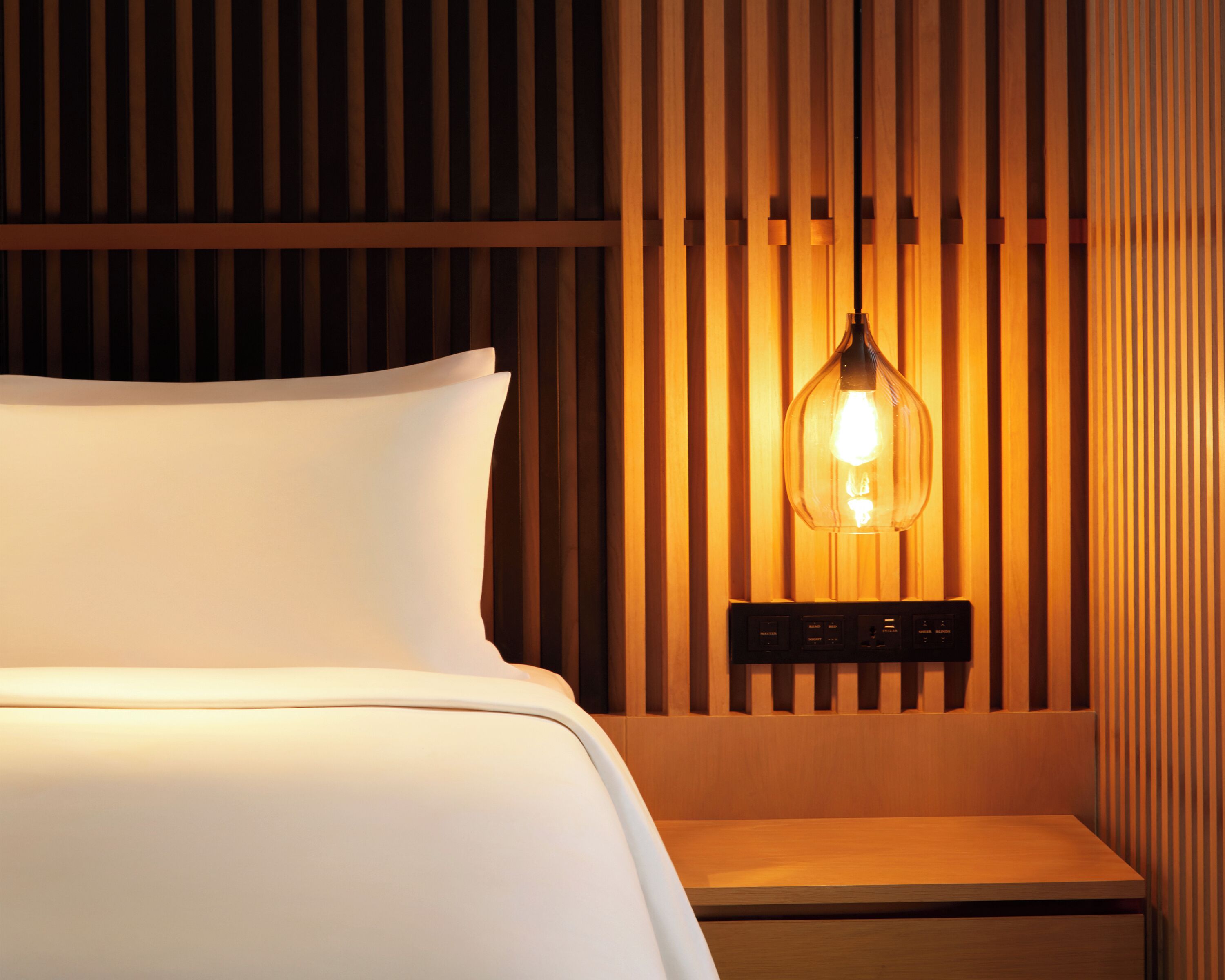 Un lit bien fait, avec une couette blanche et des oreillers, une table de chevet avec une lampe, et un mur en bois à lattes derrière le lit.