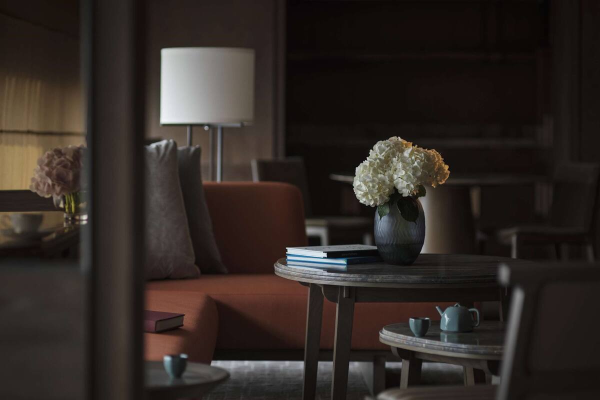 Una habitación con poca luz, un sofá, una mesa con un jarrón de flores, una lámpara y una estantería al fondo.