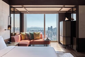A modern hotel room with a large bed, a sofa with colorful cushions, a small table, and a large window offering a city view.