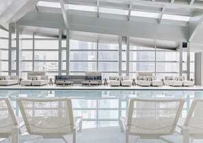 Un espace piscine intérieur moderne, doté de mobilier blanc et de grandes fenêtres offrant une vue sur la ville.