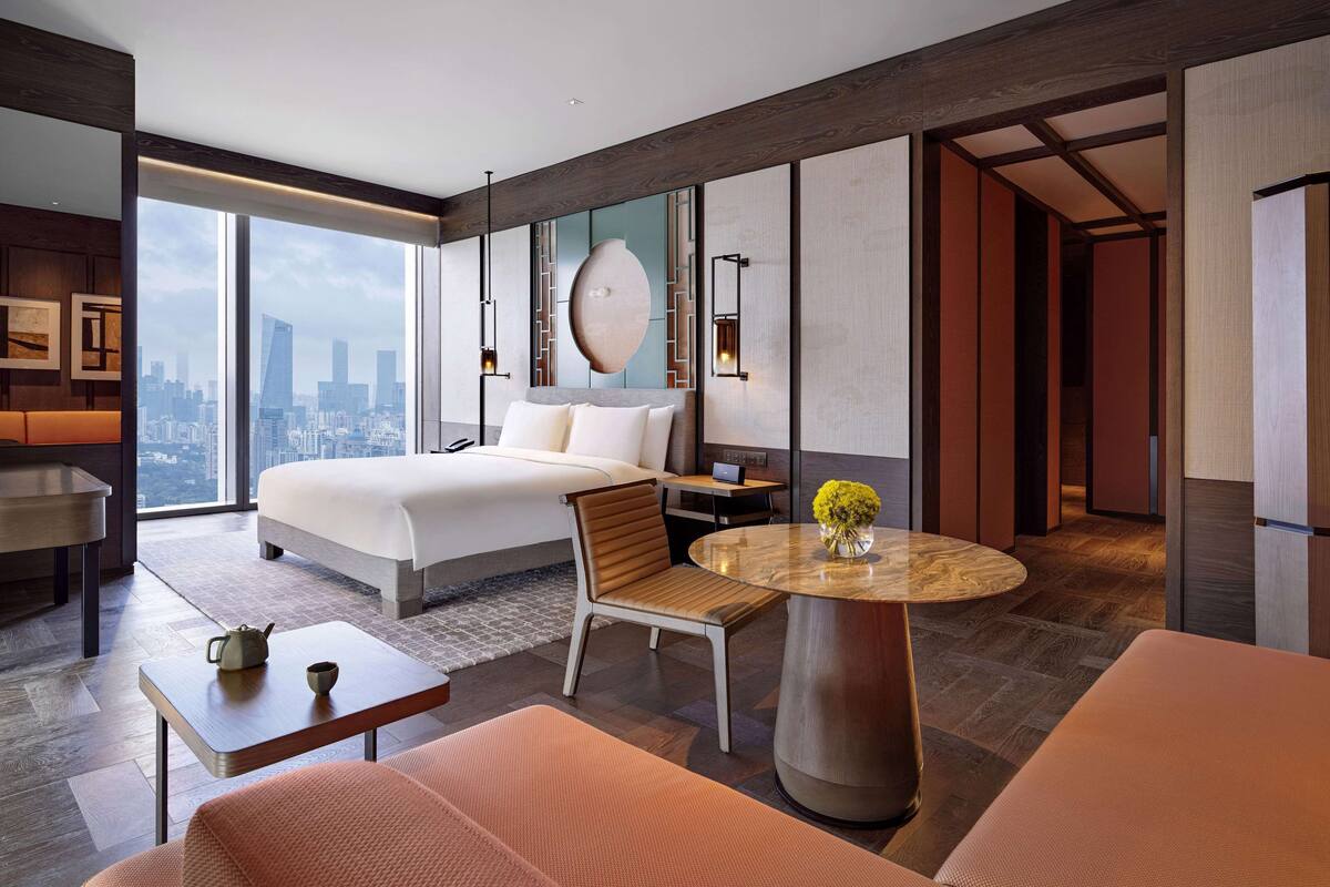 Ein modernes Hotelzimmer mit einem großen Bett, einem Essbereich, einer Sitzecke und Blick auf die Stadtlandschaft.