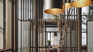 A modern dining area with high ceilings, vertical wooden slats, and a central tree. Tables are set with plates, cups, and a potted plant.