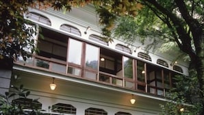 Ein zweistöckiges Gebäude mit einem Balkon im oberen Stockwerk, umgeben von viel Grün.