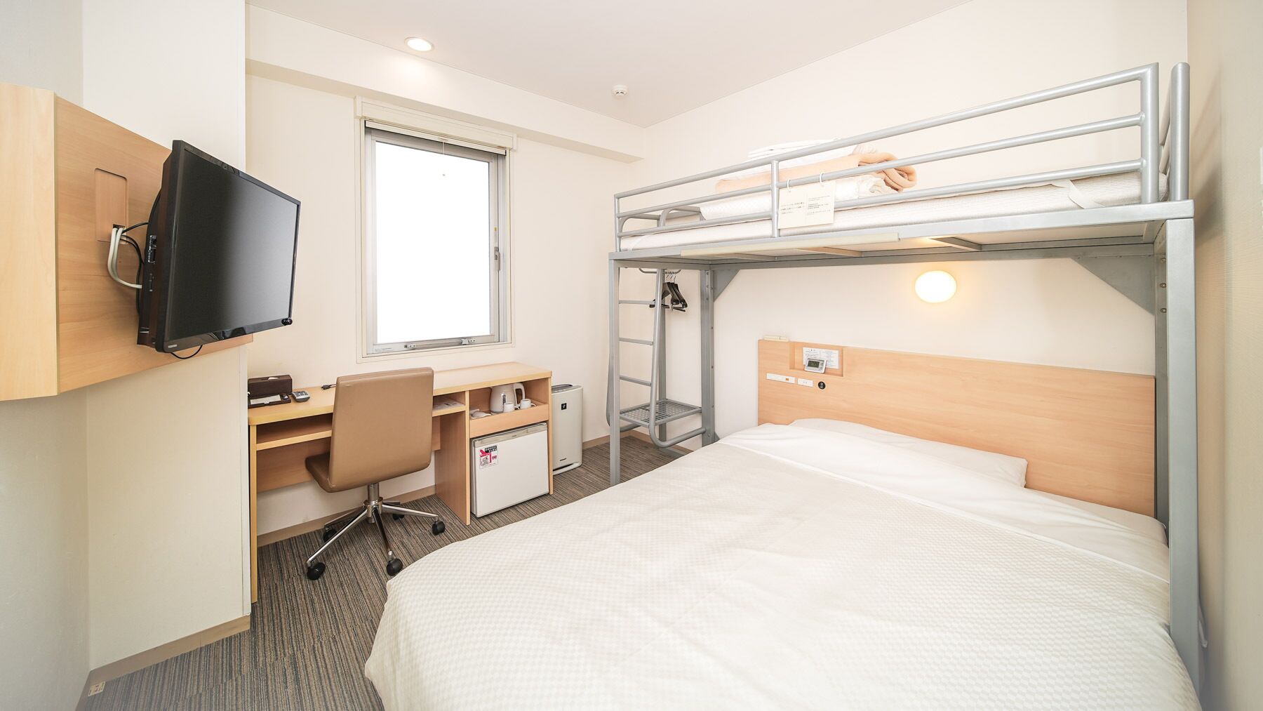 Habitación de hotel con literas, un escritorio con silla, televisión, ventana y una kitchenette pequeña.