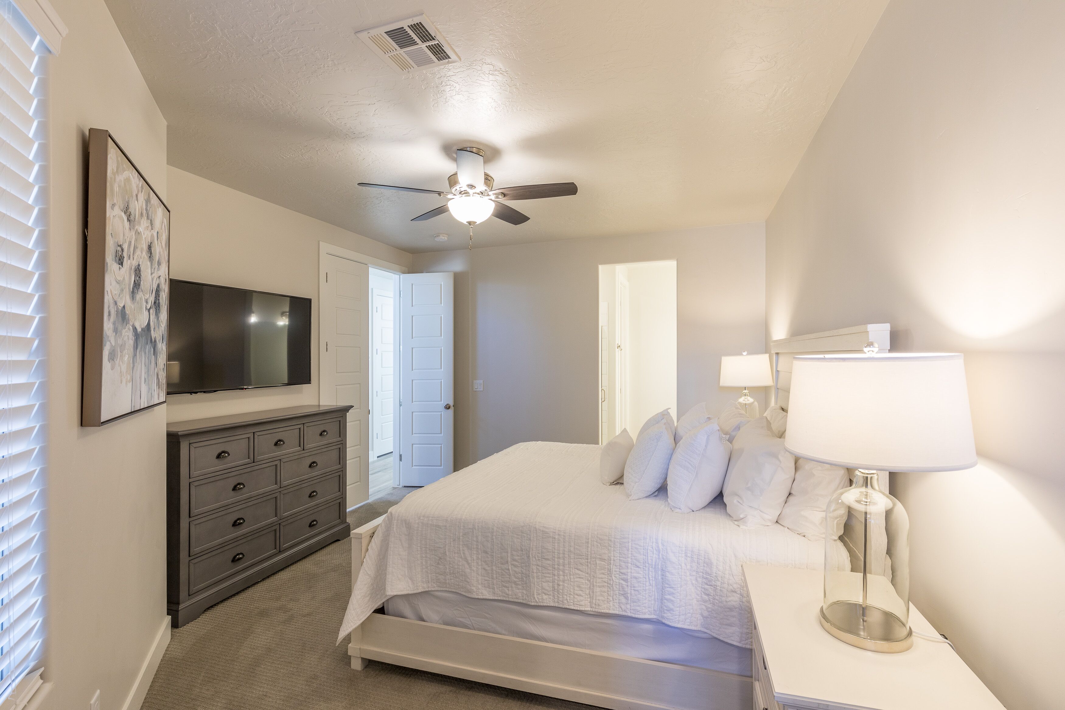 A bedroom with a bed, a dresser, a TV, a ceiling fan, and a lamp.
