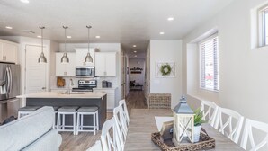 A modern kitchen with white cabinets, a central island, and pendant lighting. A dining area with a long table and white chairs is visible. - Modern 3-King 4BR Sleeps 13 - Rooftop Deck (St. George)
