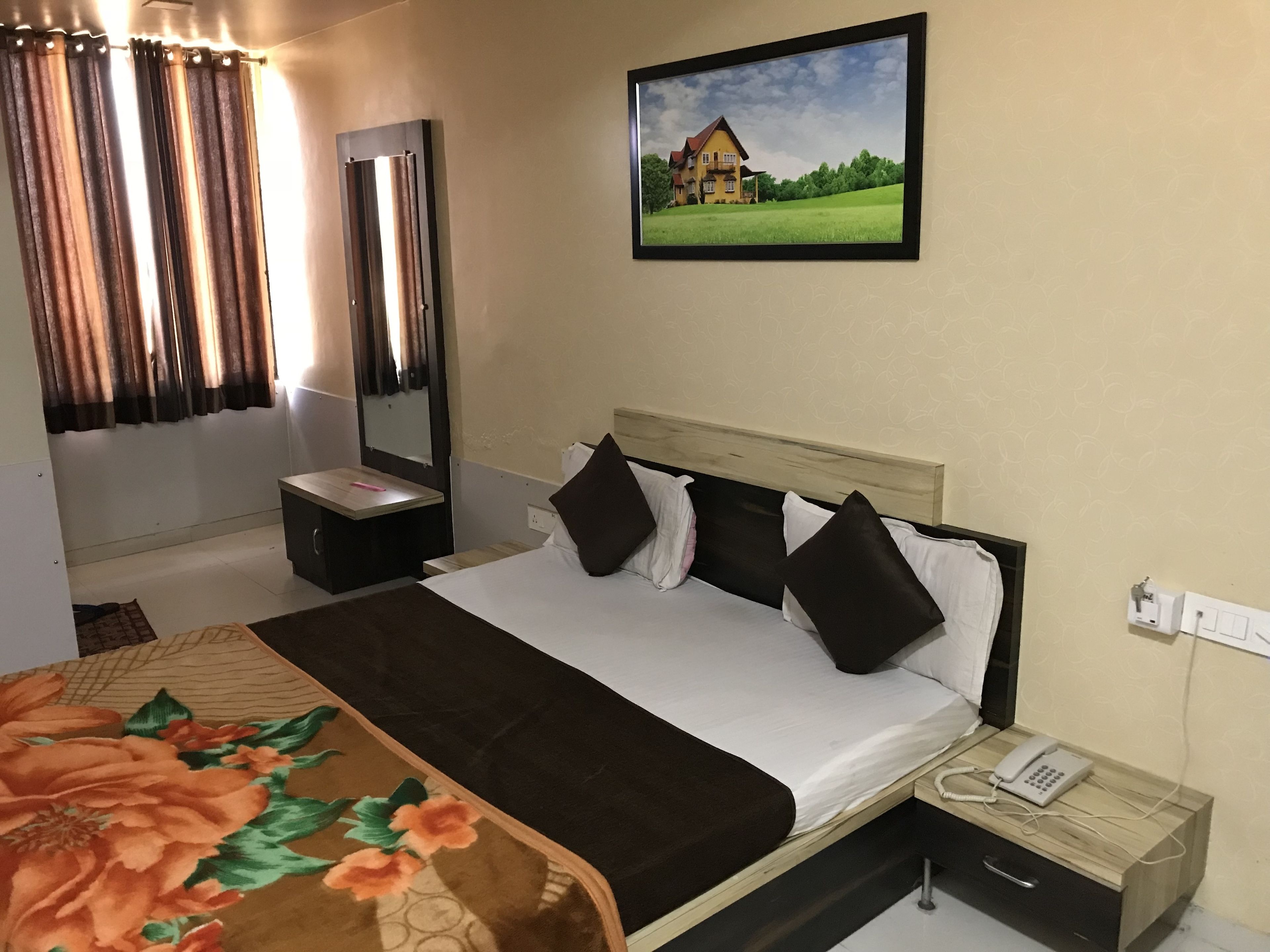 Una habitación de hotel con una cama, una mesita de noche, un teléfono y un cuadro de una casa en la pared.