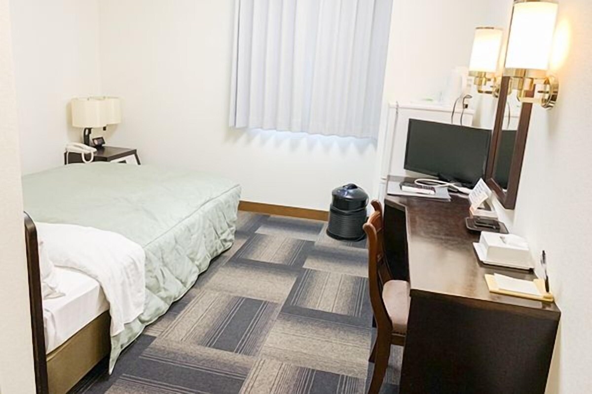 Ein Hotelzimmer mit einem Bett, einem Schreibtisch mit Computer, einem Stuhl, einer Lampe, einem Papierkorb und einem Fenster mit Jalousien.