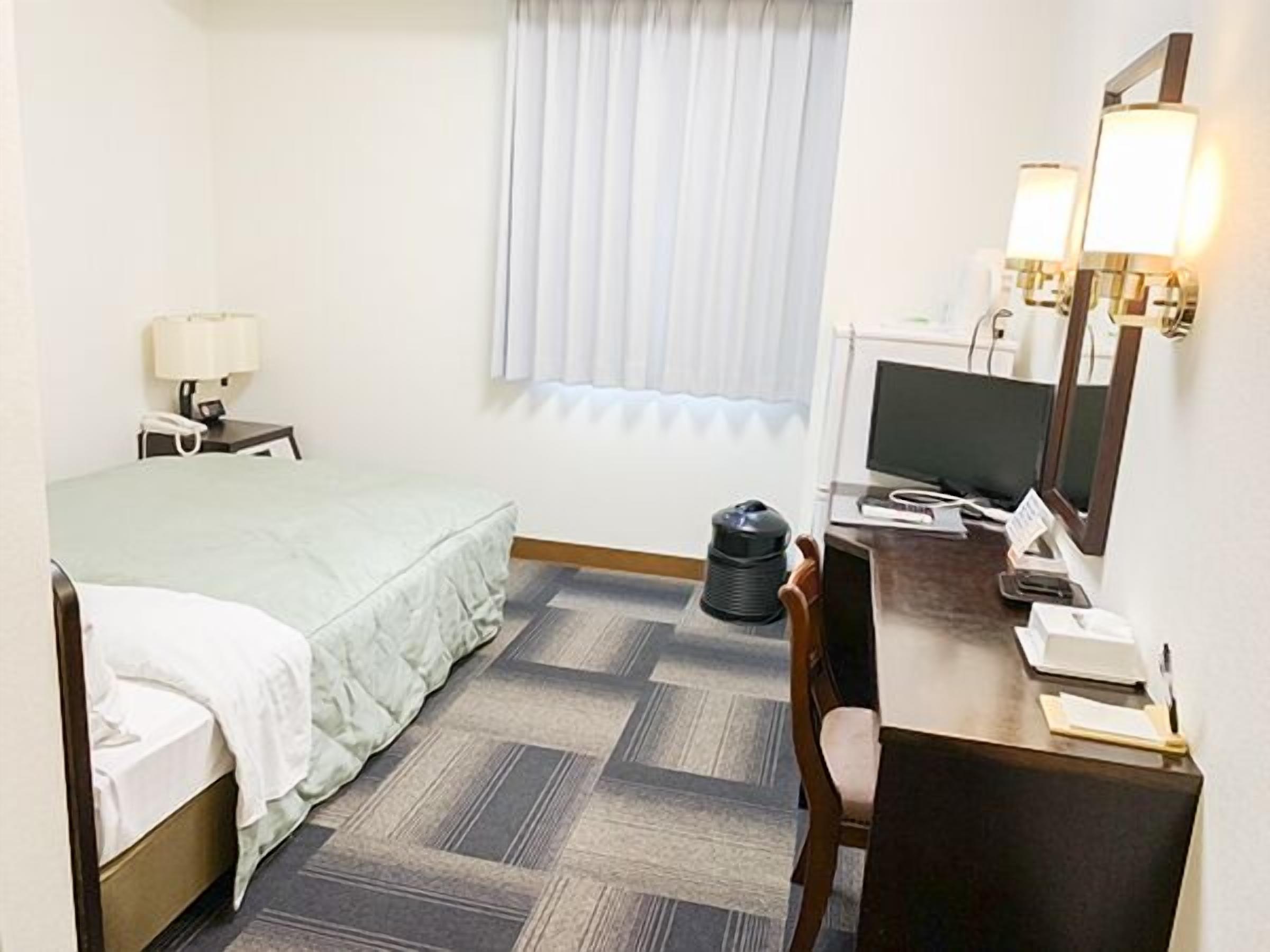 Una habitación de hotel con una cama, un escritorio con computadora, una silla, una lámpara, un cubo de basura y una ventana con persianas.