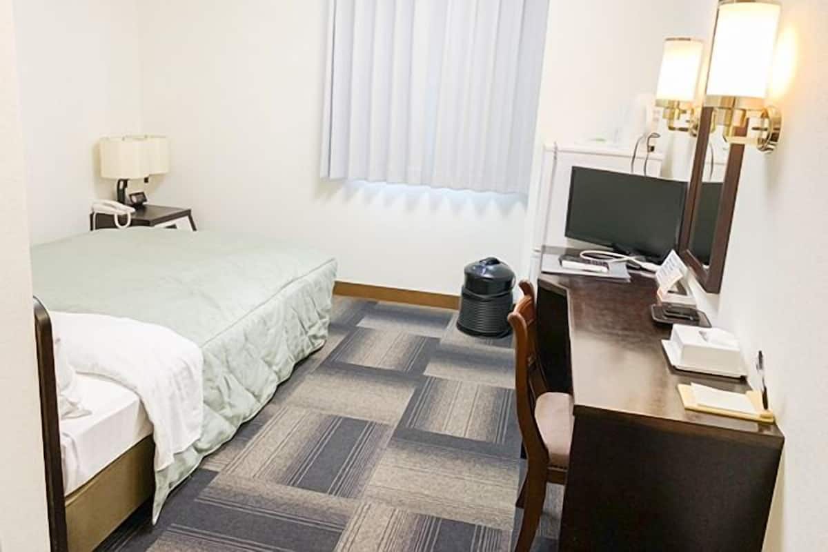 A hotel room with a bed, a desk with a computer, a chair, a lamp, a trash can, and a window with blinds.