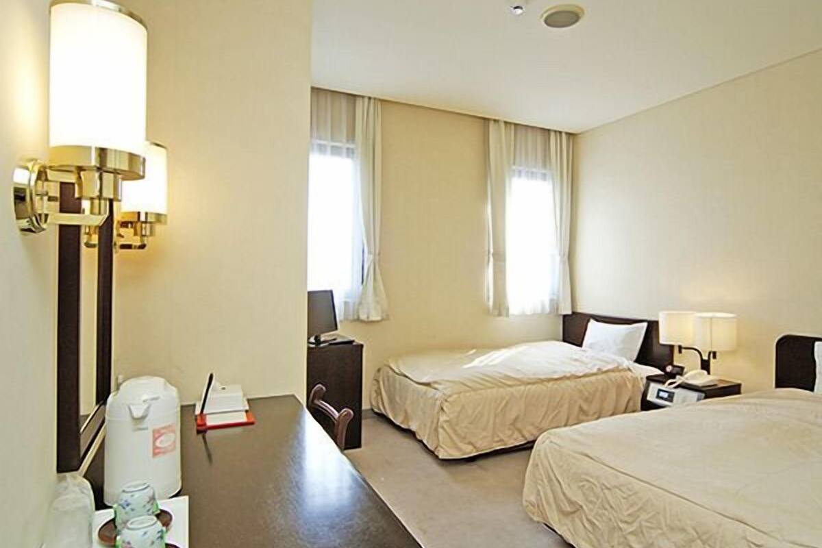 A hotel room with two beds, a desk, a chair, a lamp, a nightstand, and a window with curtains.
