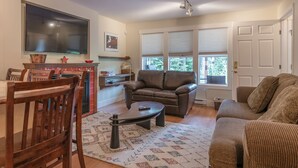 Ein Wohnzimmer mit braunem Ledersofa, einem Holzesstisch mit Stühlen, einem an der Wand befestigten Flachbildfernseher und einem Fenster mit Jalousien.