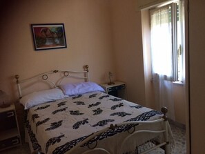 A bedroom with a bed, a nightstand, a bedside table, a window with curtains, and a painting on the wall.