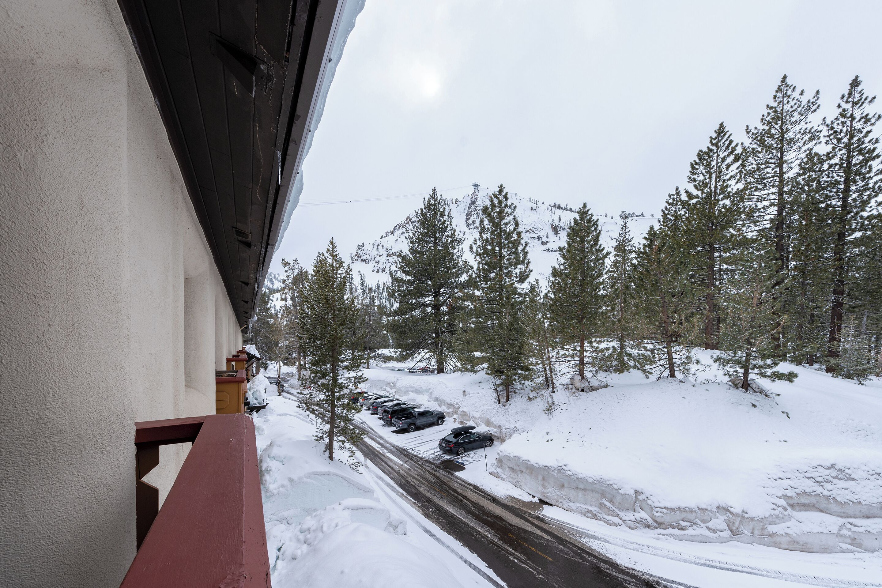 A snow-covered road with parked cars and a view of a forested mountain area.
