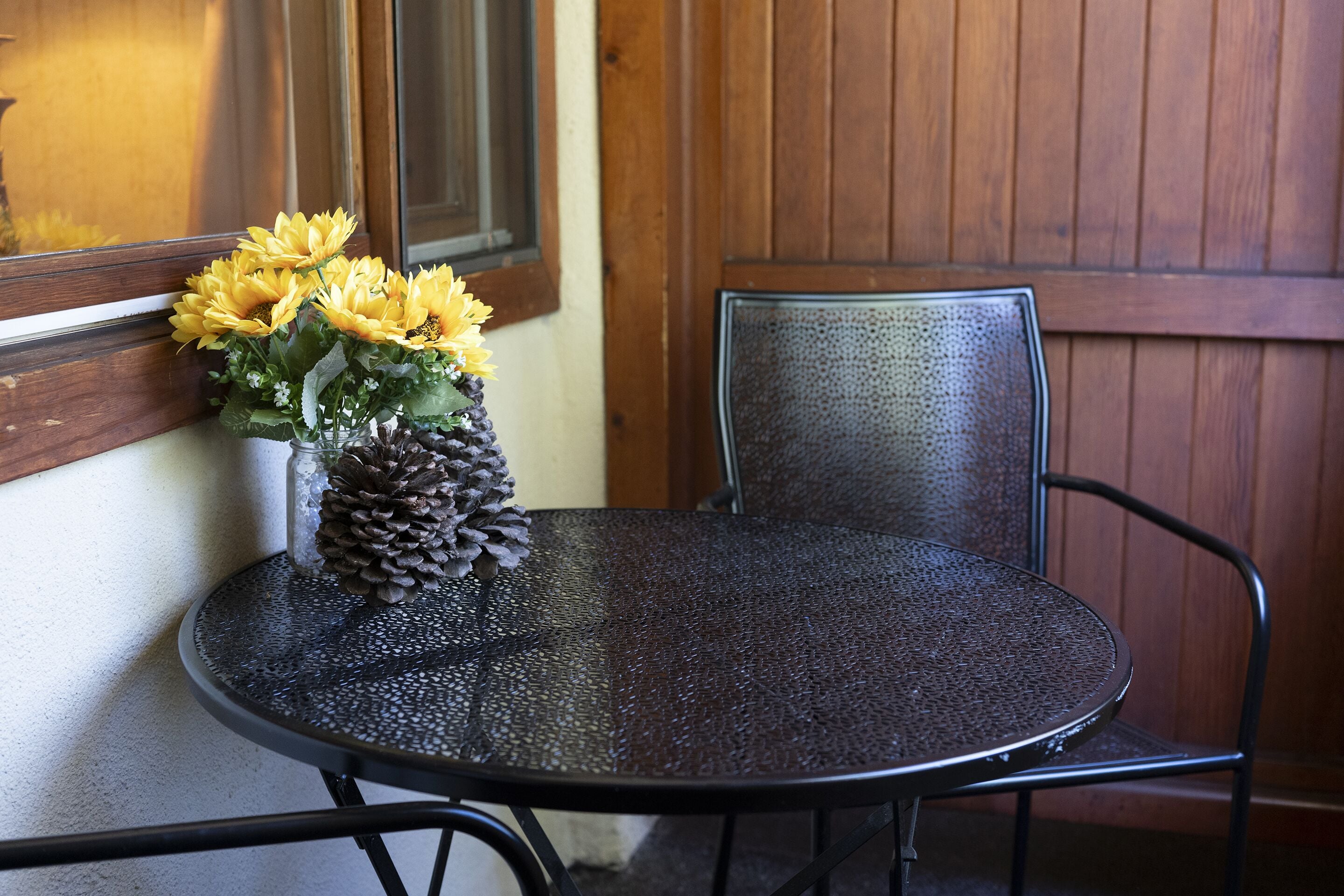 A round metal table with a metal chair, a vase with yellow flowers and a pine cone, and a wooden paneled wall in the background.
