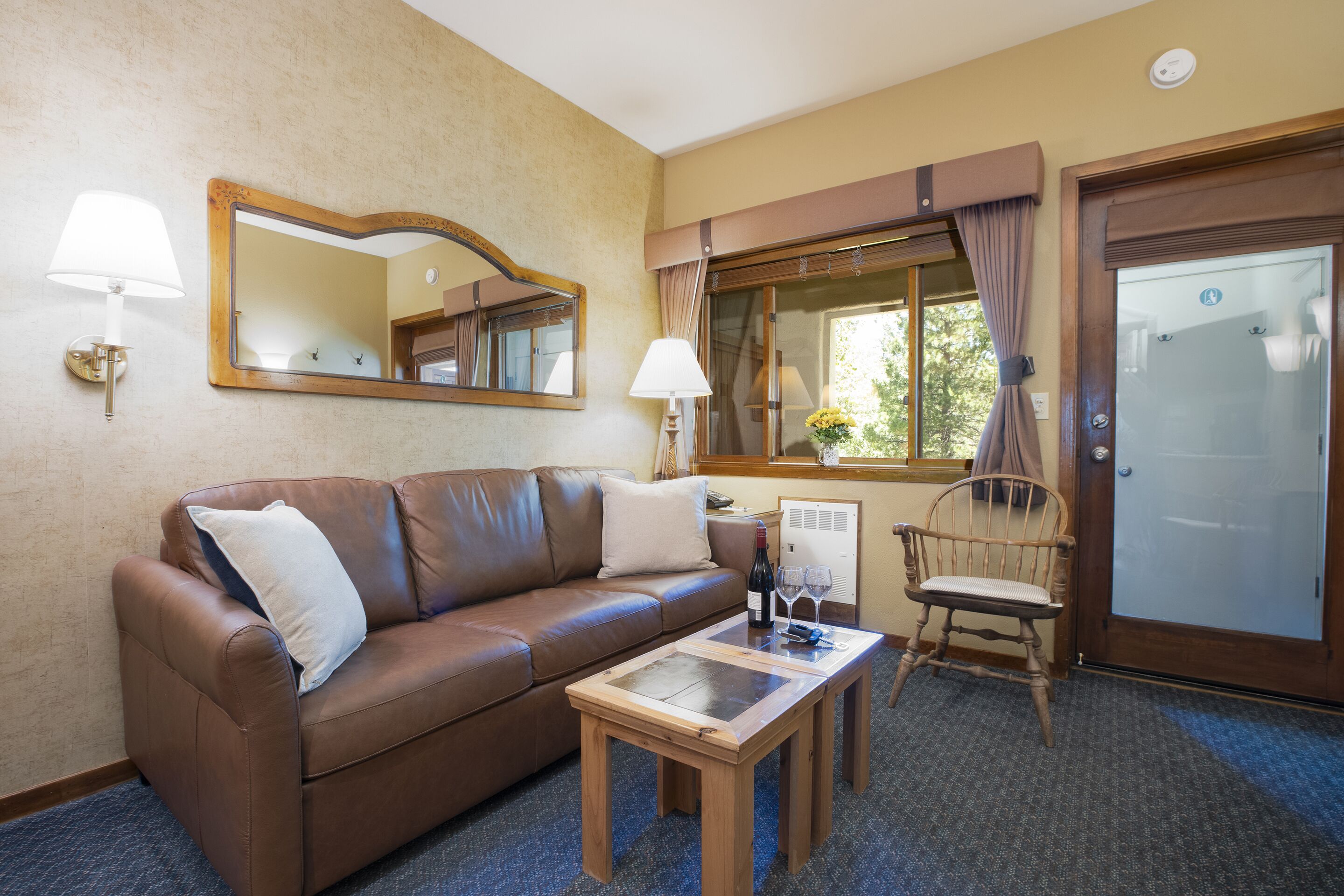 Una habitación con un sofá de cuero marrón, una mesa de centro de madera, una silla de madera, un espejo, una ventana con cortinas y una puerta.