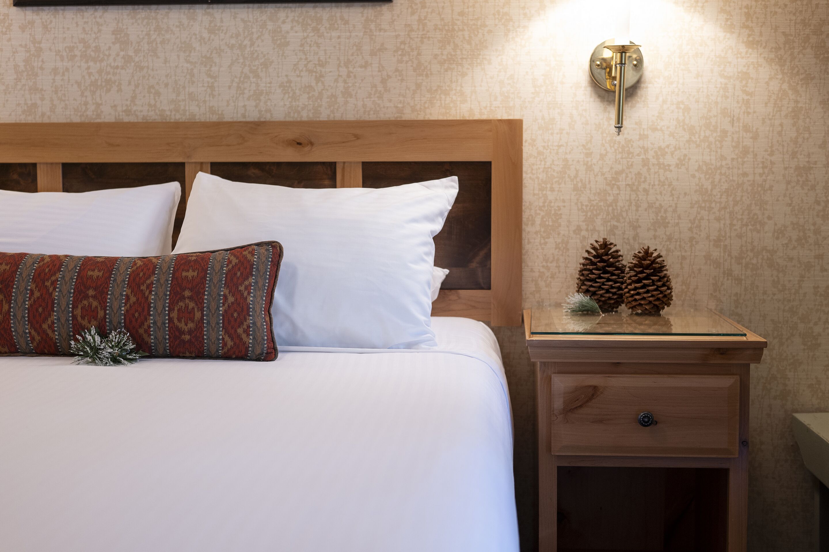 Un lit bien fait, avec du linge de lit blanc et un coussin à motifs, une table de chevet en bois avec un tiroir et un luminaire fixé au mur.
