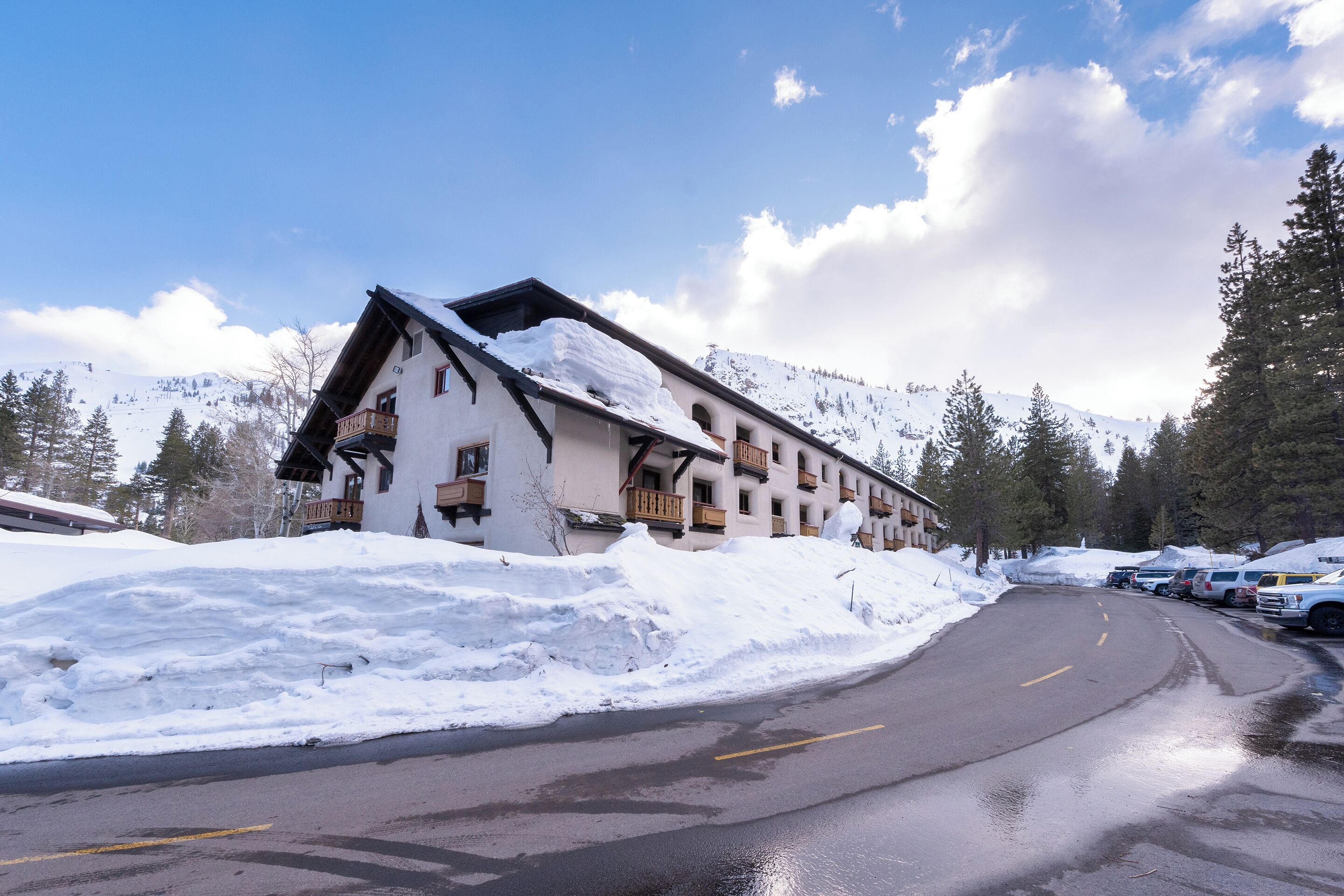 Un edificio cubierto de nieve con tejado inclinado y balcones de madera, ubicado junto a una carretera con vehículos estacionados.