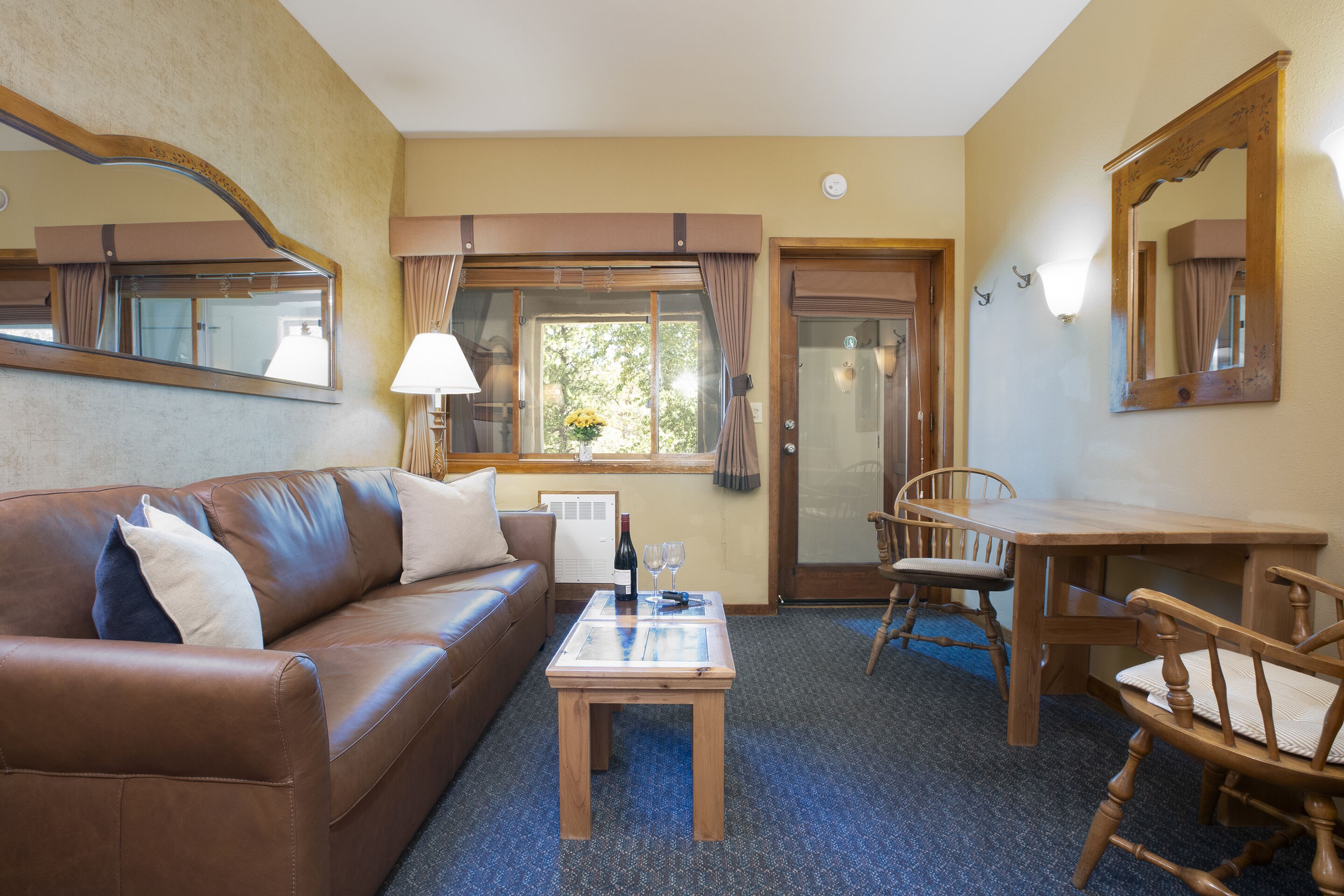 A furnished room with a brown leather sofa, a wooden coffee table, a dining table with chairs, and a large mirror.