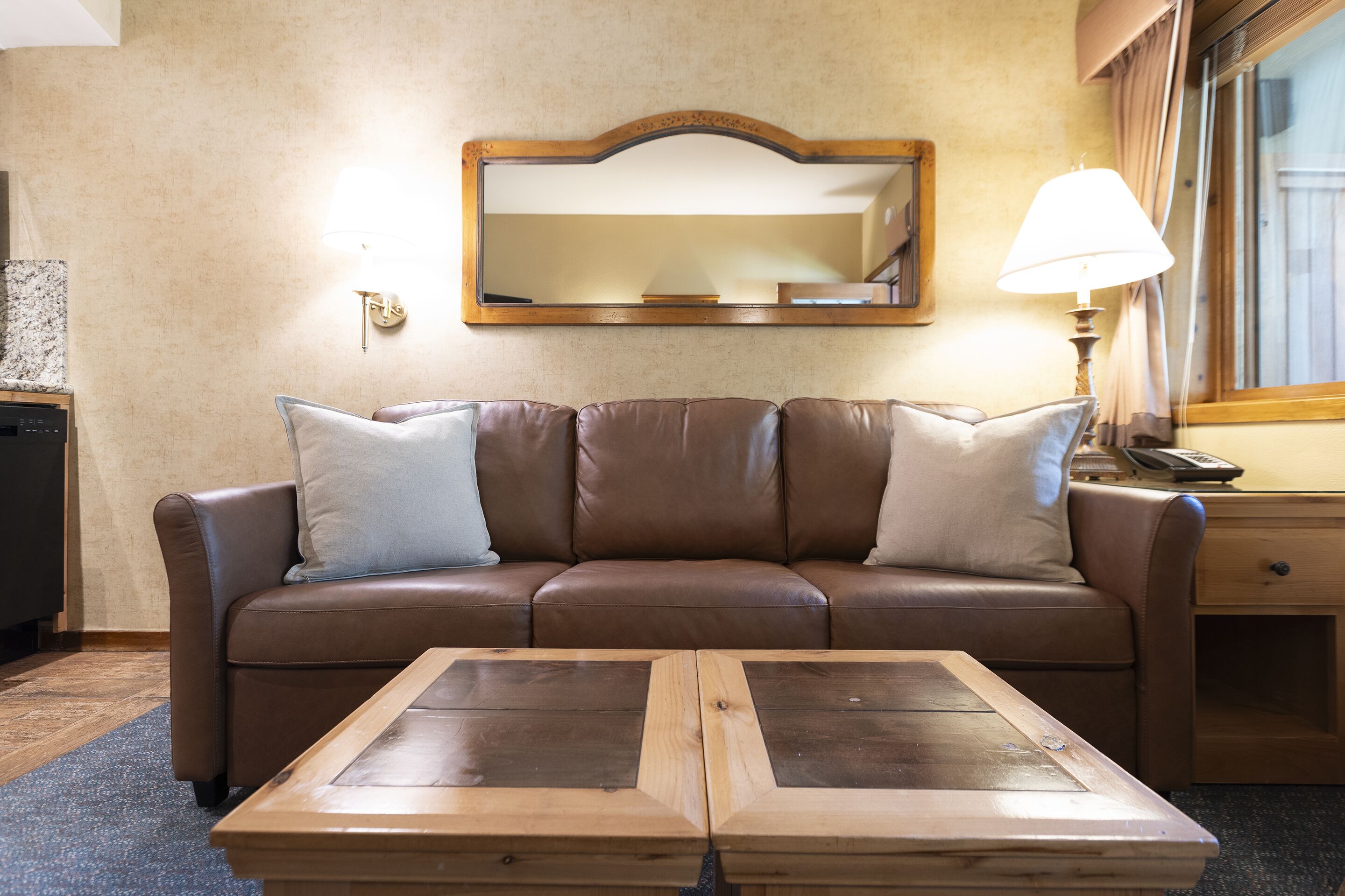 A brown leather sofa with two pillows, a wooden coffee table, a lamp, and a mirror on the wall.