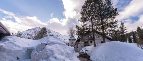 A snow-covered landscape with a building, a large tree, and mountains in the background.