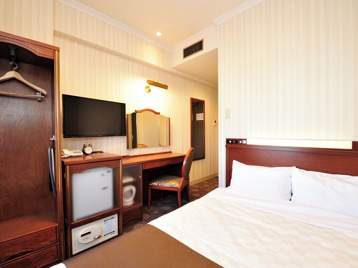 A hotel room with a bed, a desk with a chair, a television, a mirror, and a wardrobe.