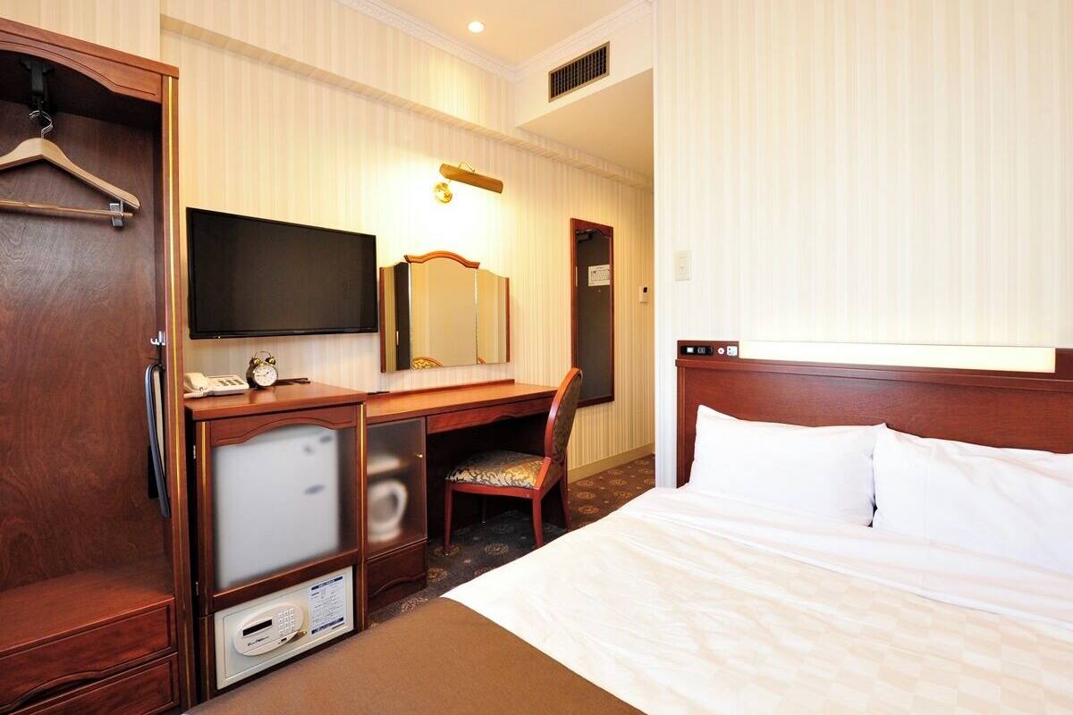 A hotel room with a bed, a desk with a chair, a television, a mirror, and a wardrobe.