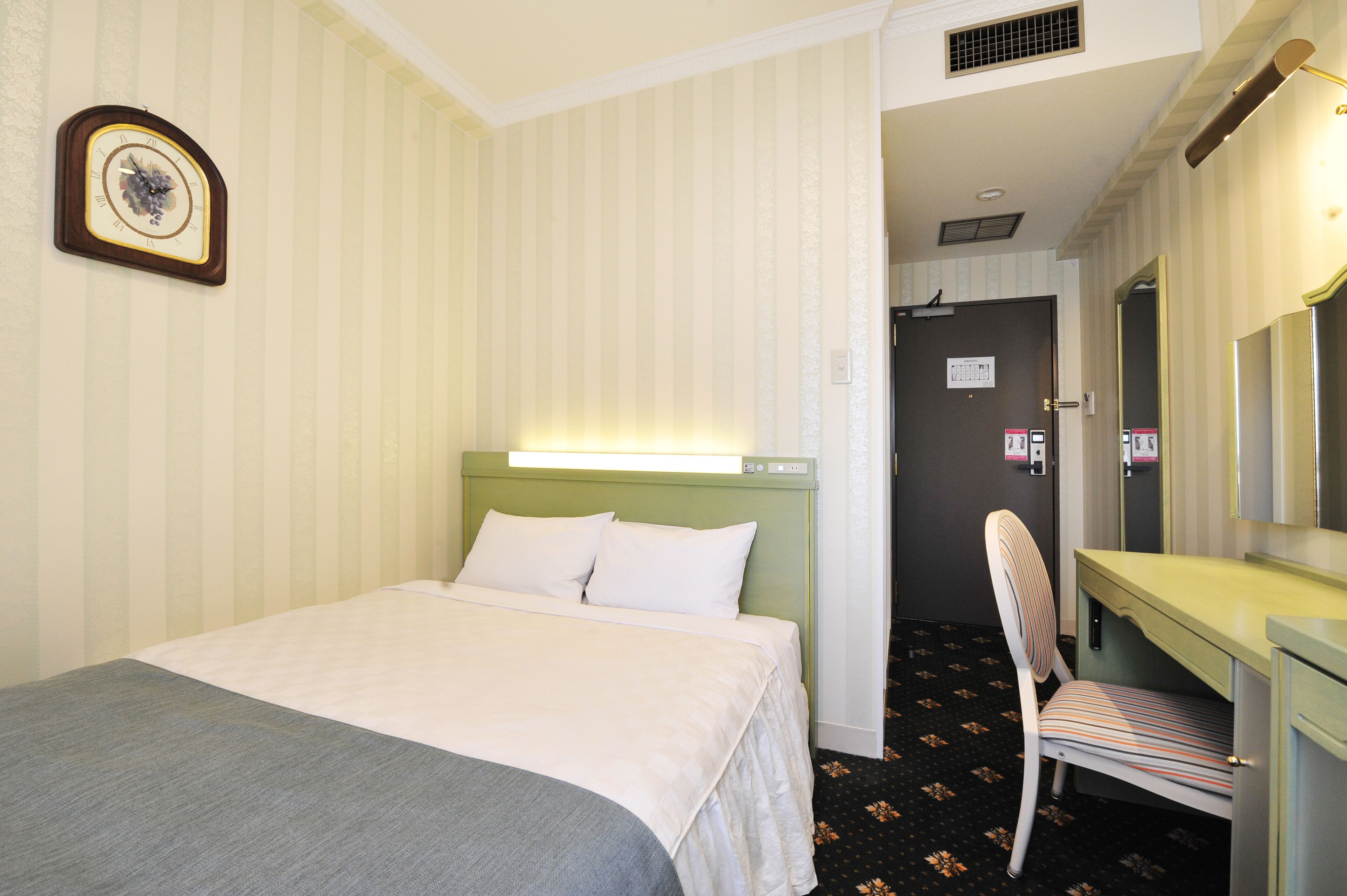 A hotel room with a bed, a desk, a chair, a clock, and a door leading to another room.