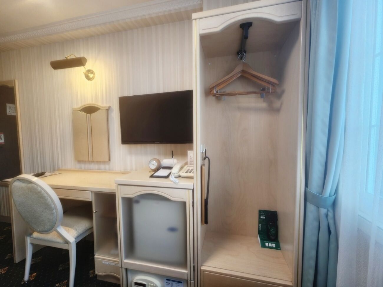 A hotel room with a desk, chair, wardrobe, and a hanging coat rack.