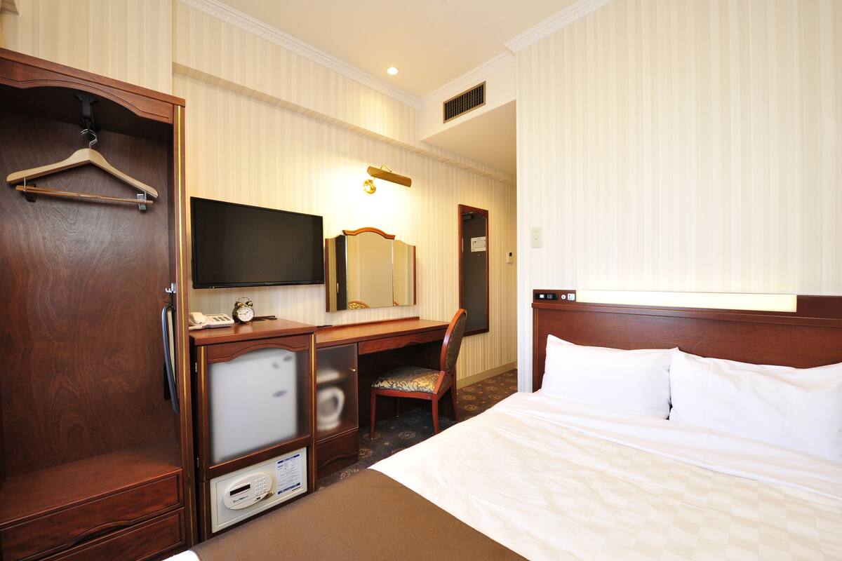 A hotel room with a bed, a desk with a computer, a chair, a TV, and a wardrobe.