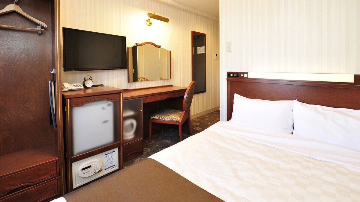 A hotel room with a bed, a desk with a chair, a television, a mirror, and a wardrobe.