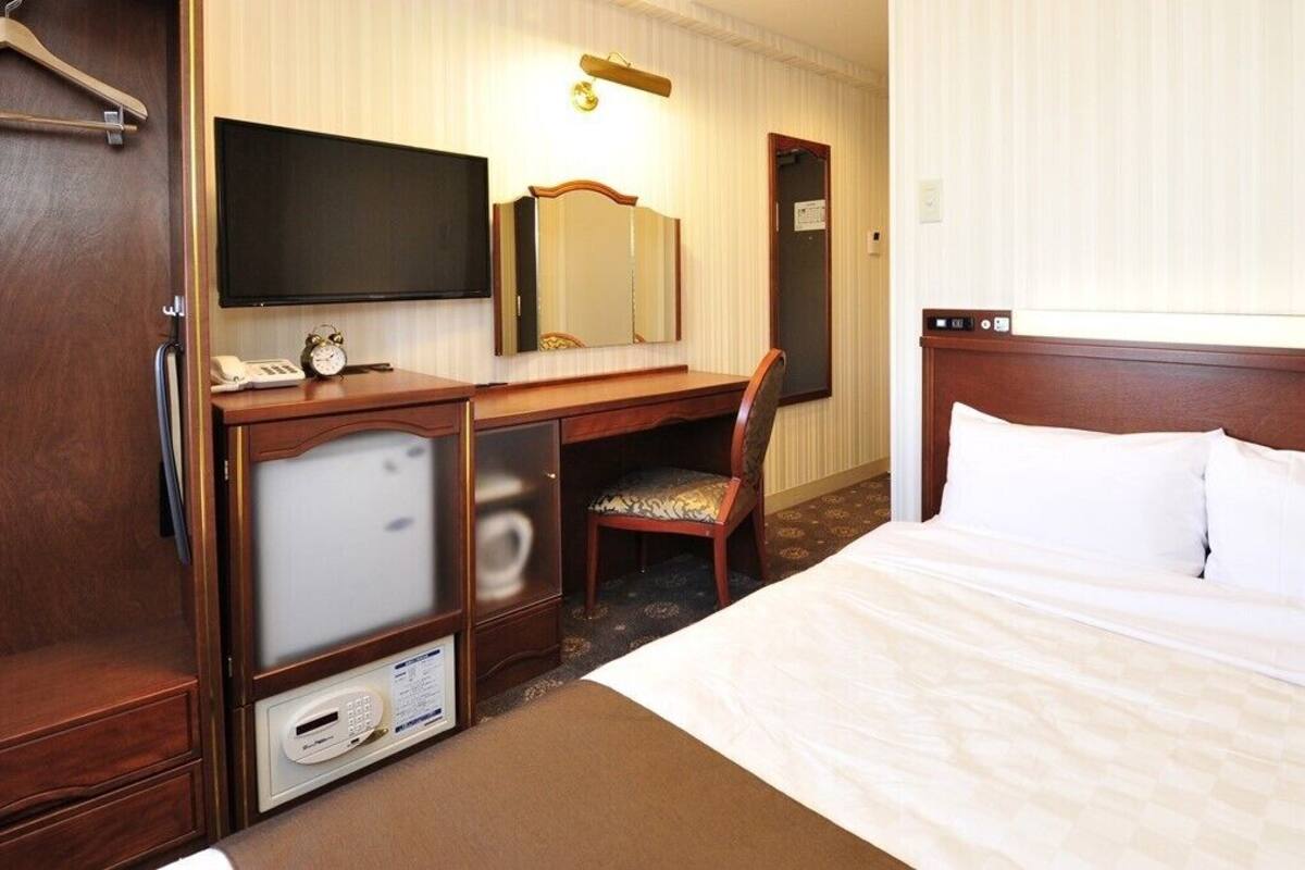 A hotel room with a bed, a desk with a chair, a television, a mirror, and a wardrobe.