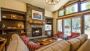 A cozy living room with a stone fireplace, comfortable seating, and a large window offering a view of the outdoors. - Sunriver Luxury Home at Caldera Springs - TRAI94 (Sunriver)