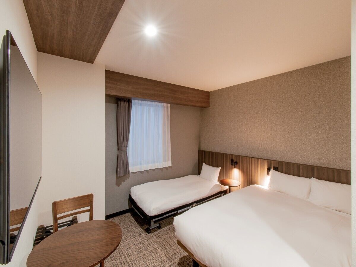 A hotel room with two beds, a desk, a chair, a window with curtains, and a ceiling light.