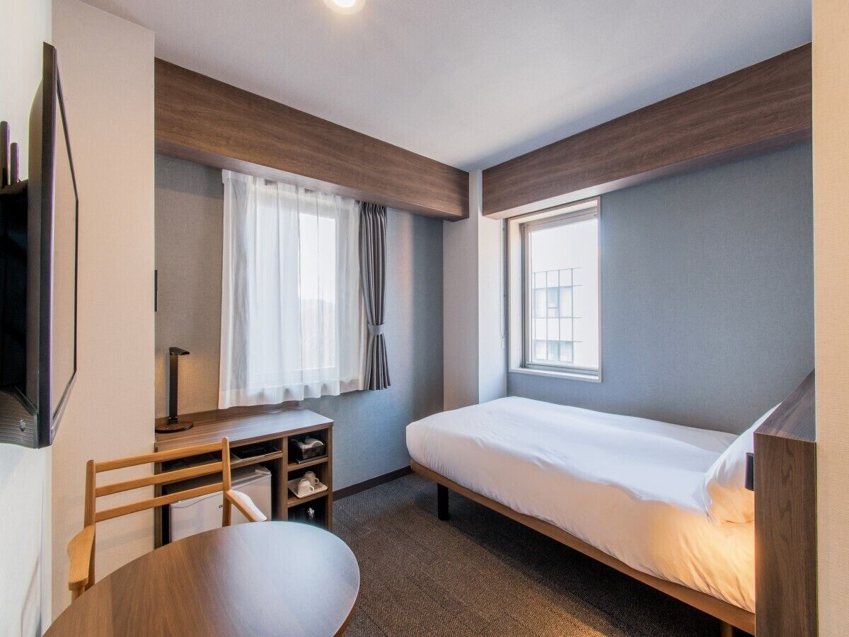 A compact hotel room with a single bed, a desk with a chair, a small table, and a window with curtains.