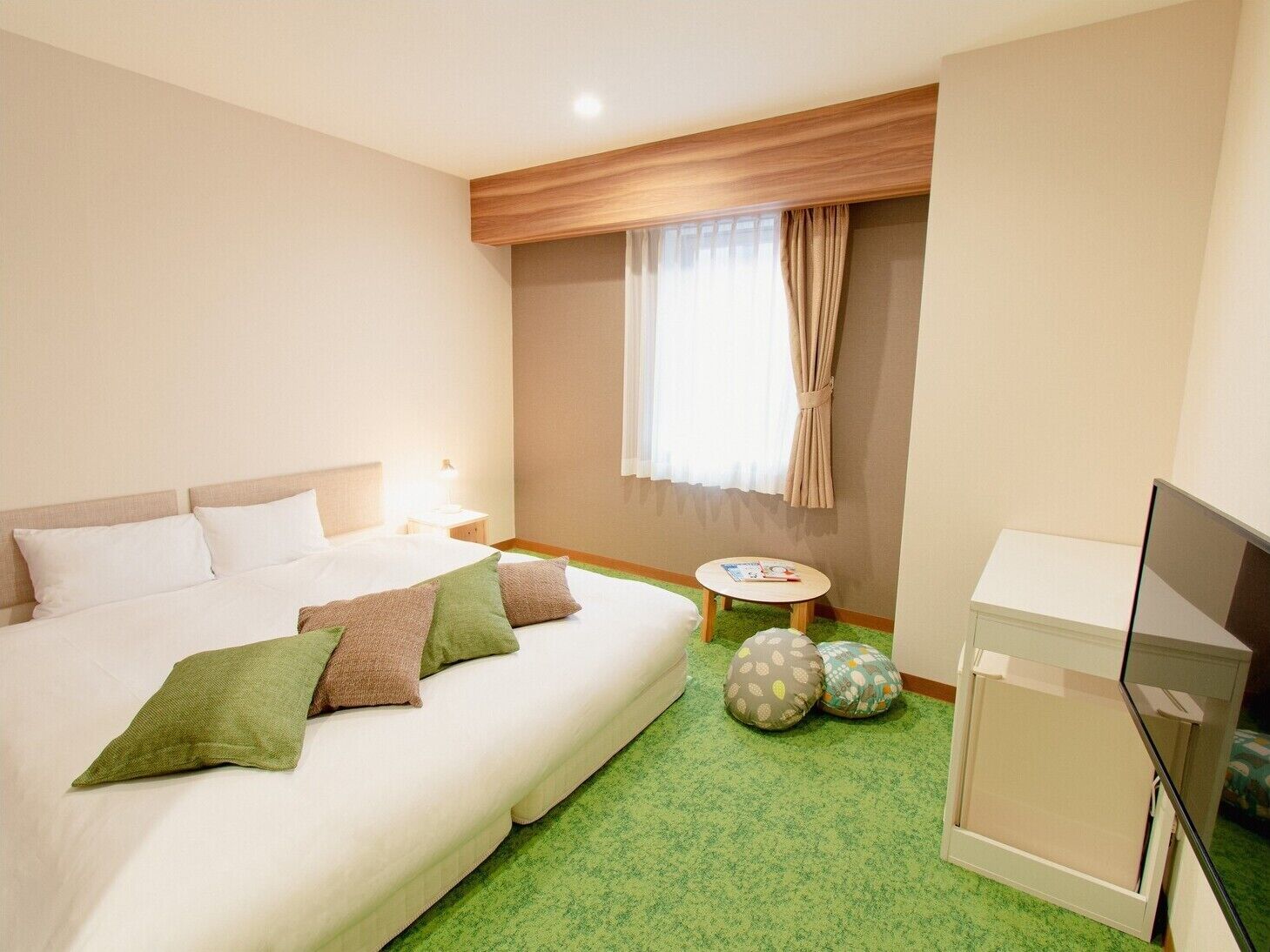 A hotel room with a bed, a small table with a book, a TV, and a window with curtains.