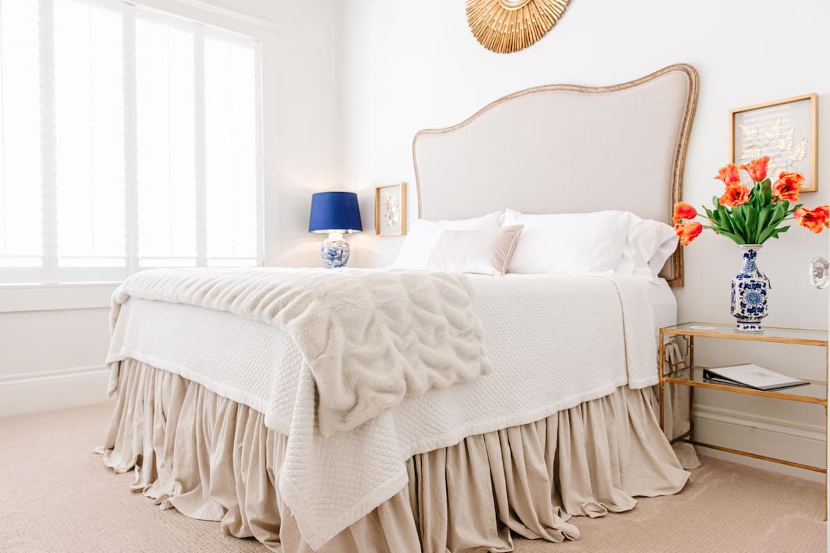 Una cama bien tendida con una colcha blanca y una manta color beige, una lámpara azul y un florero con flores anaranjadas sobre una mesita de noche.