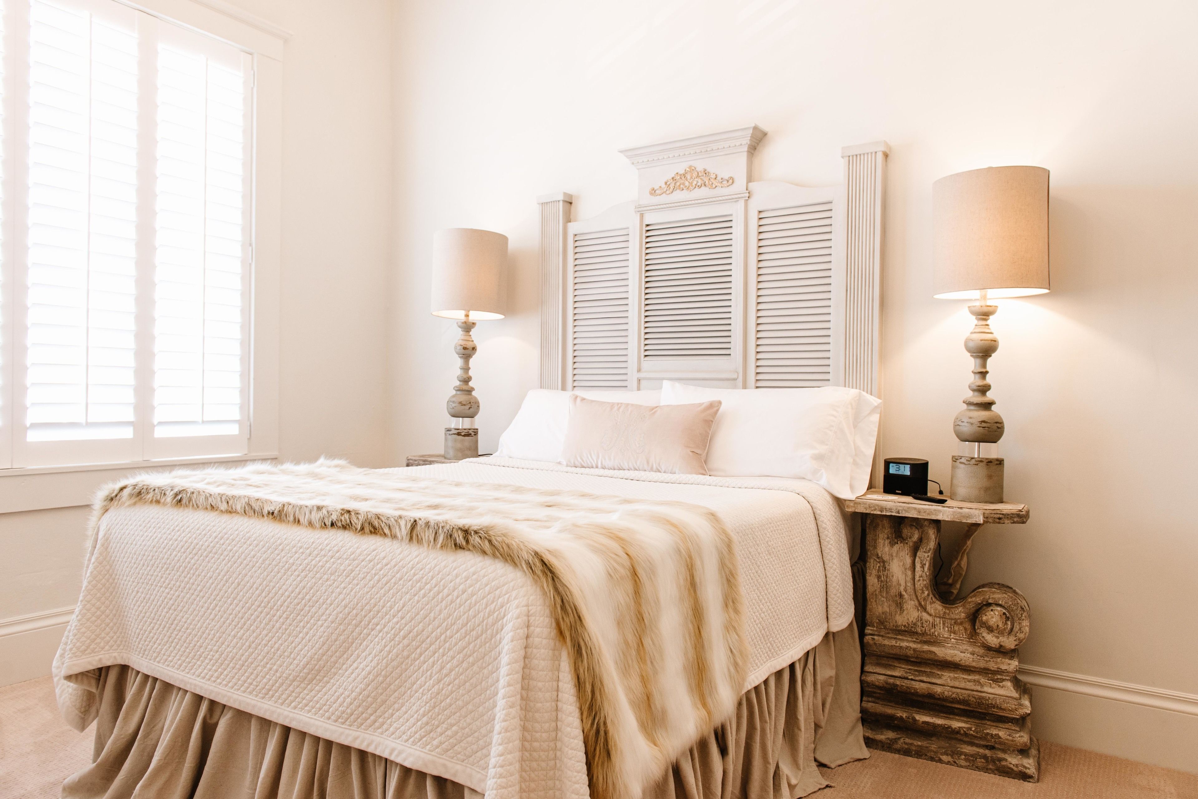 A bedroom with a bed, bedside lamps, a nightstand, a window with blinds, and a decorative wall piece above the bed.