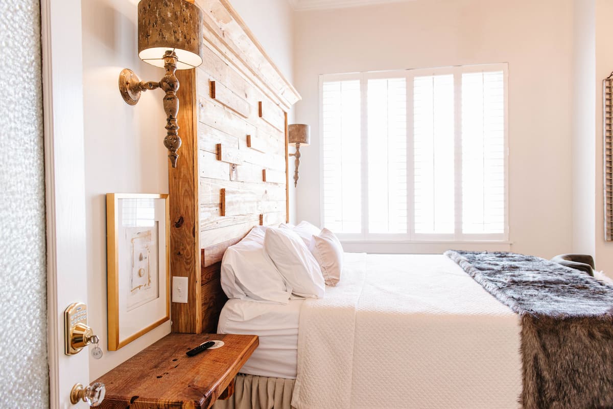 Un dormitorio con cabecera de madera, una mesita de noche, cama con ropa de cama blanca y una ventana con persianas.