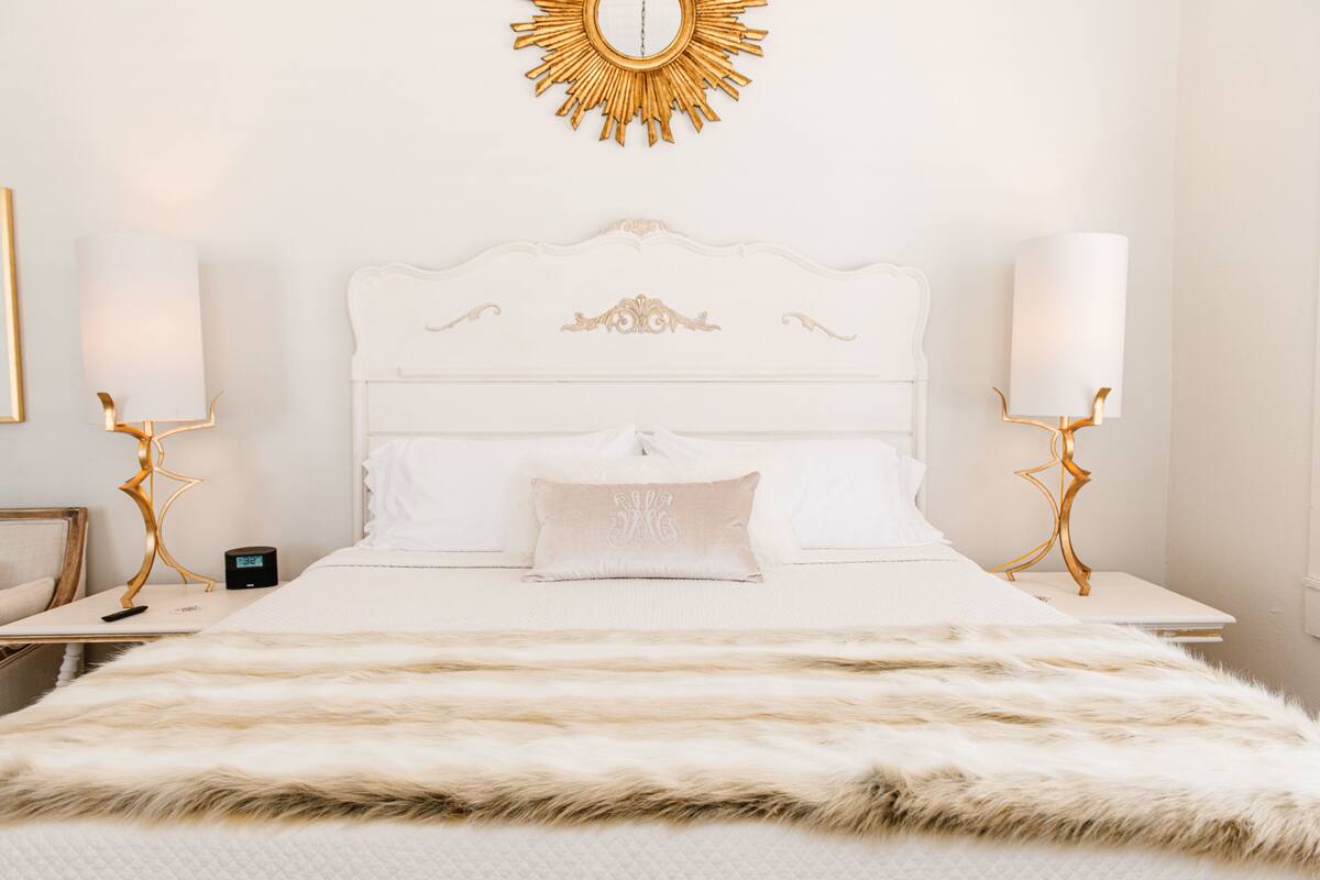 Una cama blanca con un edredón blanco mullido, un espejo con marco dorado en la pared, y dos mesitas de noche con lámparas.