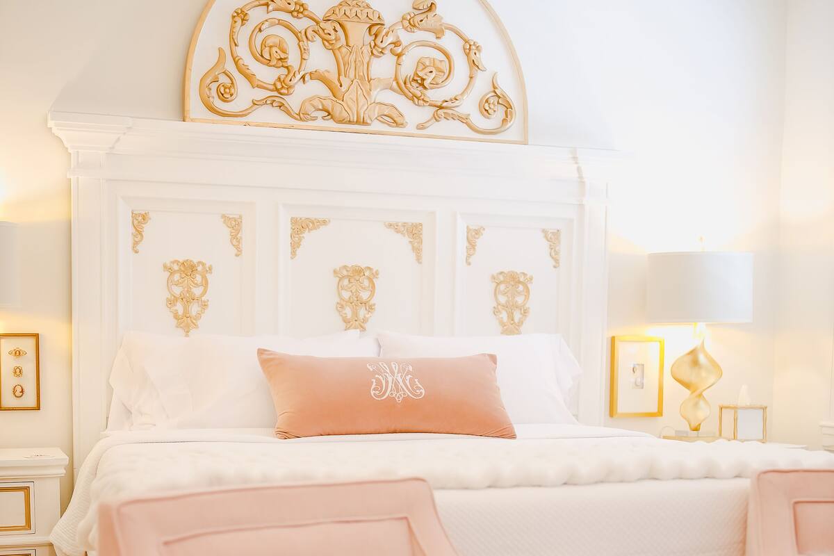 Un dormitorio con una cama grande, un cabecero decorativo, un sillón rosa, una mesita redonda y una mesita de noche con una lámpara.