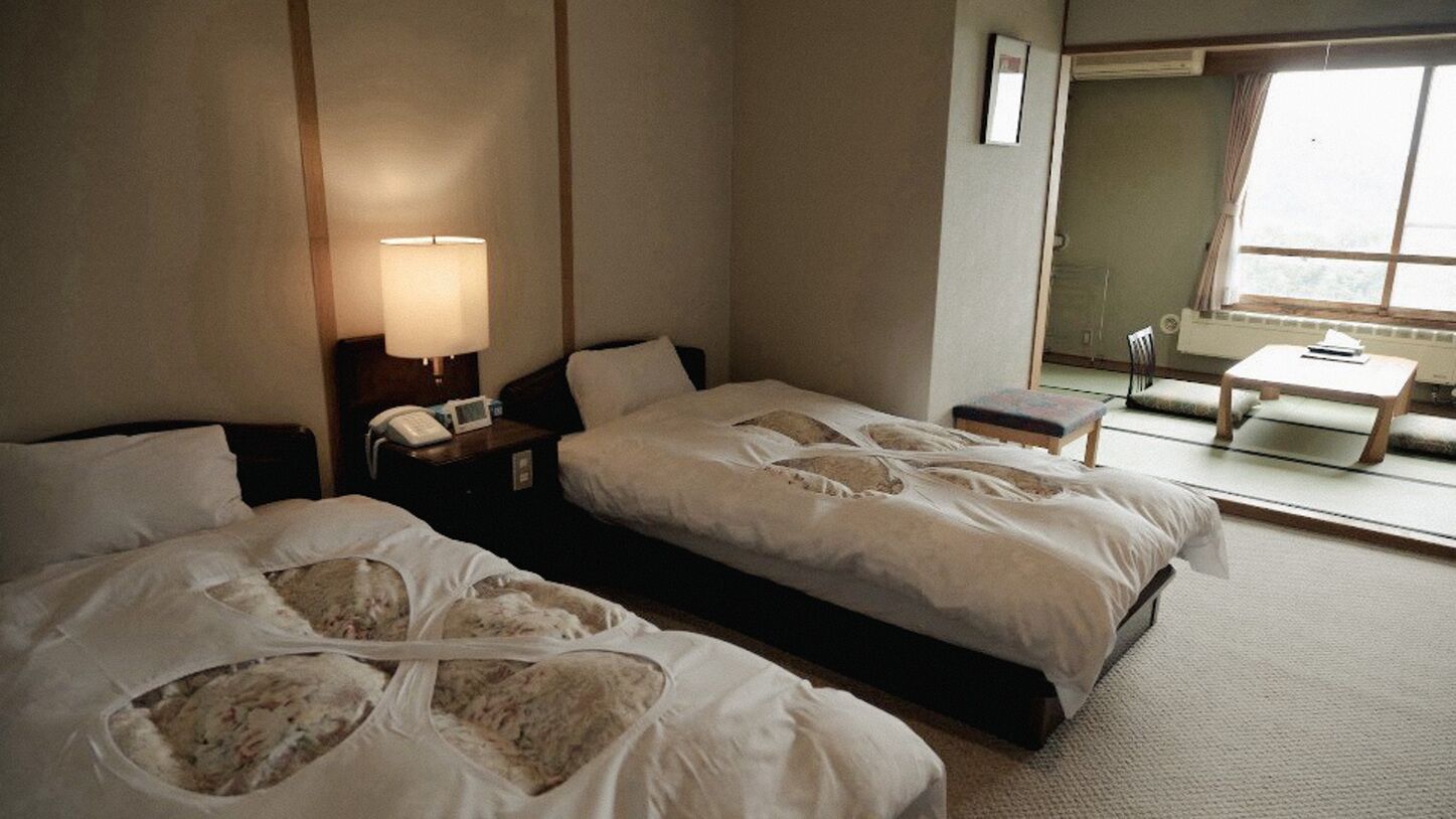 Habitación de hotel con dos camas, una mesita de noche con lámpara, una pequeña mesa con una silla y una ventana con vistas.