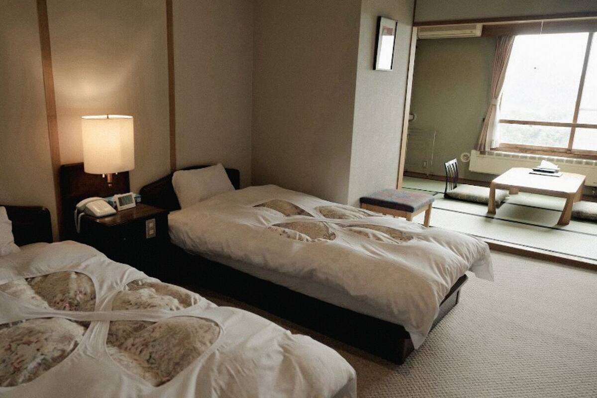 A hotel room with two beds, a bedside table with a lamp, a small table with a chair, and a window with a view.