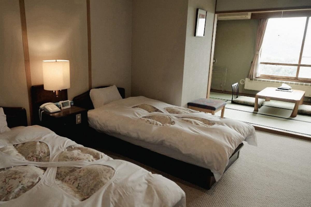 Habitación de hotel con dos camas, una mesita de noche con lámpara, una pequeña mesa con una silla y una ventana con vistas.
