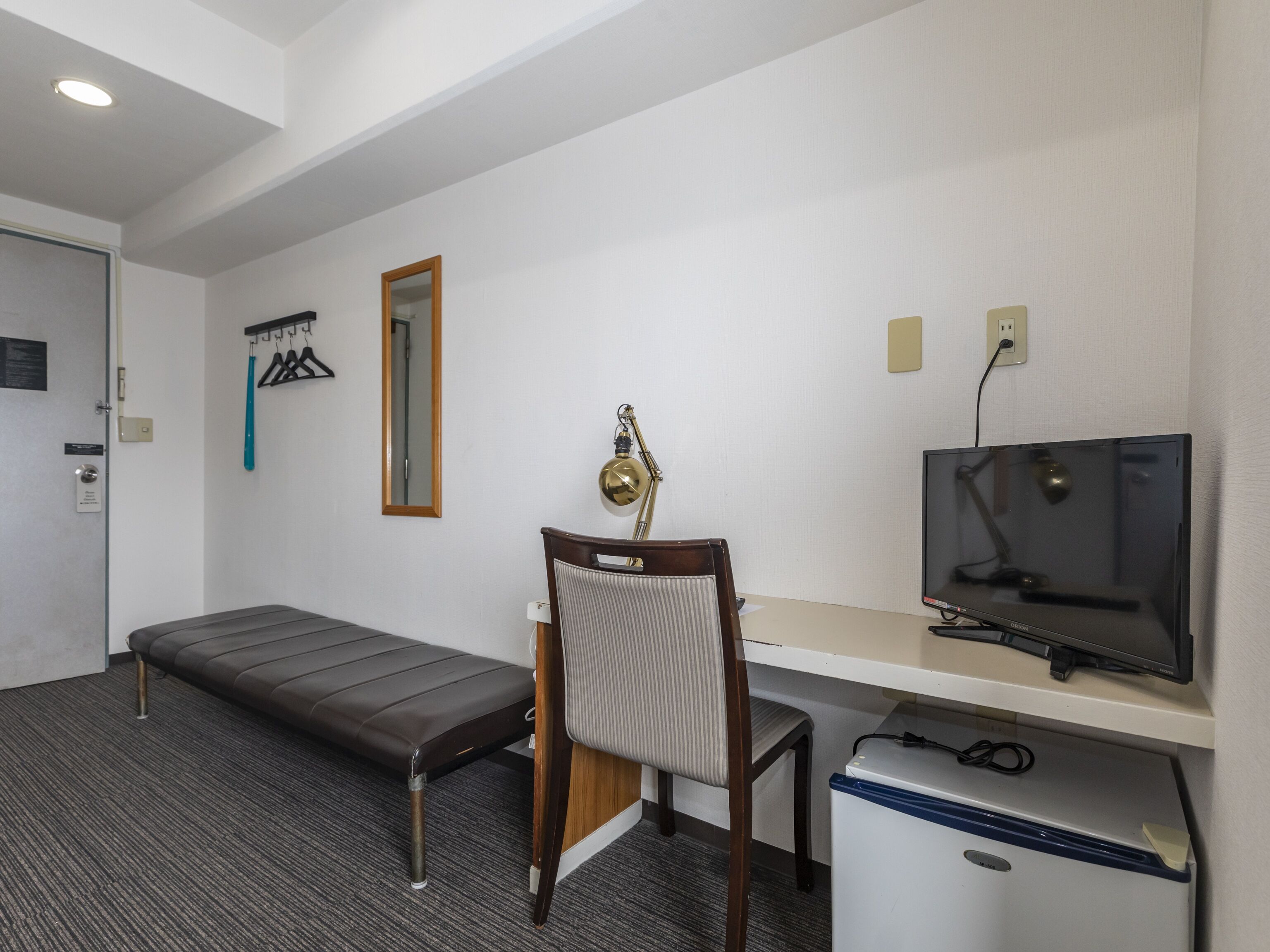 A hotel room with a bed, a desk with a computer and TV, a chair, and a small refrigerator.