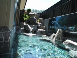 Piscina cristalina con rocas y una cascada, rodeada por un edificio con un amplio ventanal de vidrio.