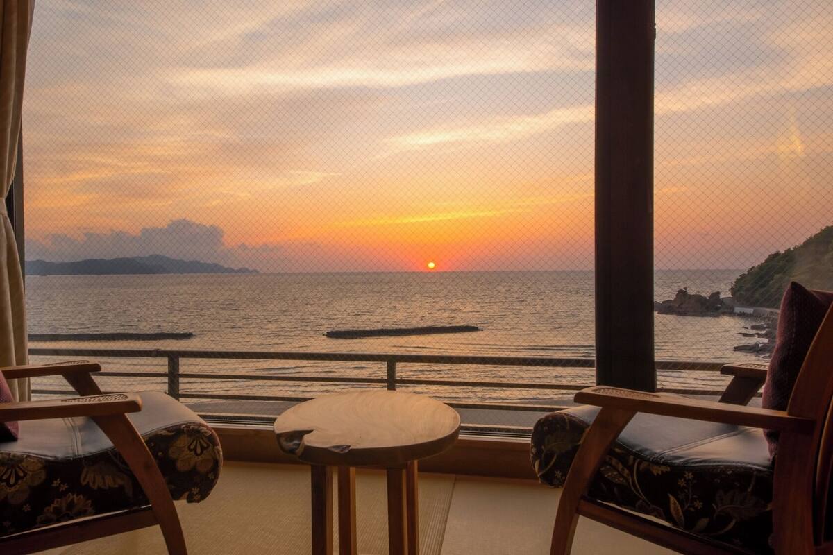 A room with two wooden chairs and a small table, overlooking a sunset over the sea.