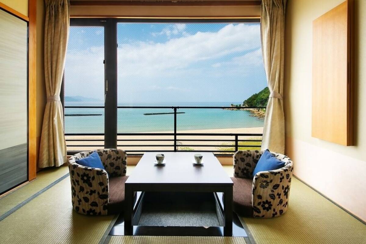 A room with tatami flooring, a sliding door, a table with two chairs, and a view of a beach and ocean.