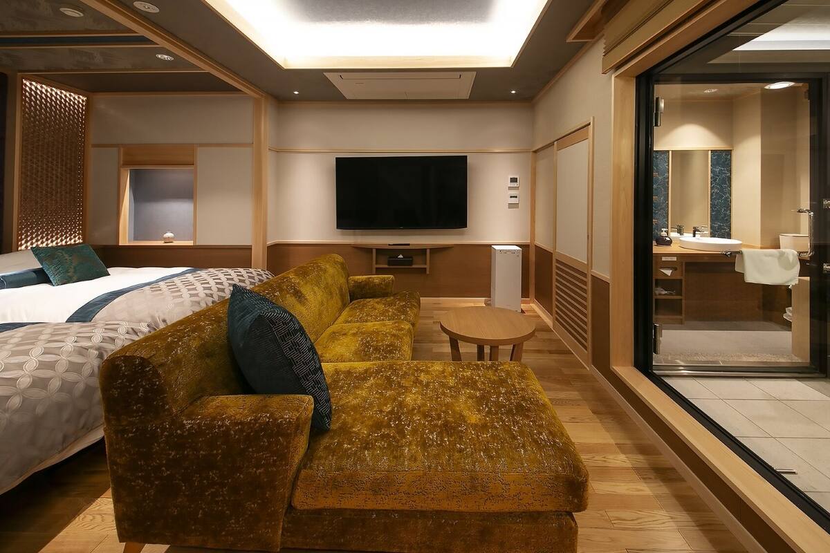 A hotel room with a bed, sofa, TV, and a bathroom with a shower and sink.